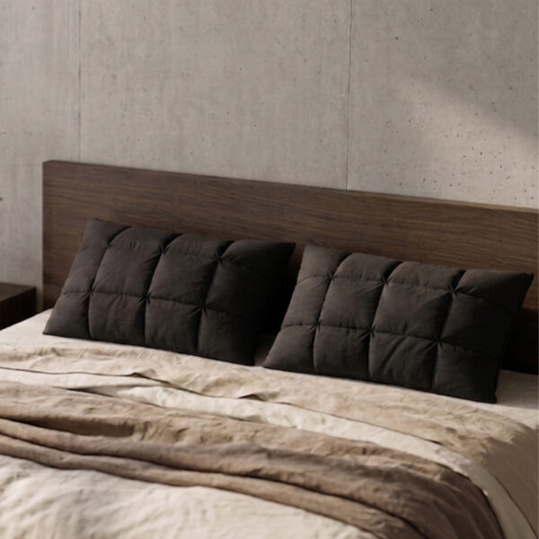Pair of premium black pillows styled on a bed in a modern minimalist bedroom with a dark wood headboard and raw concrete wall.